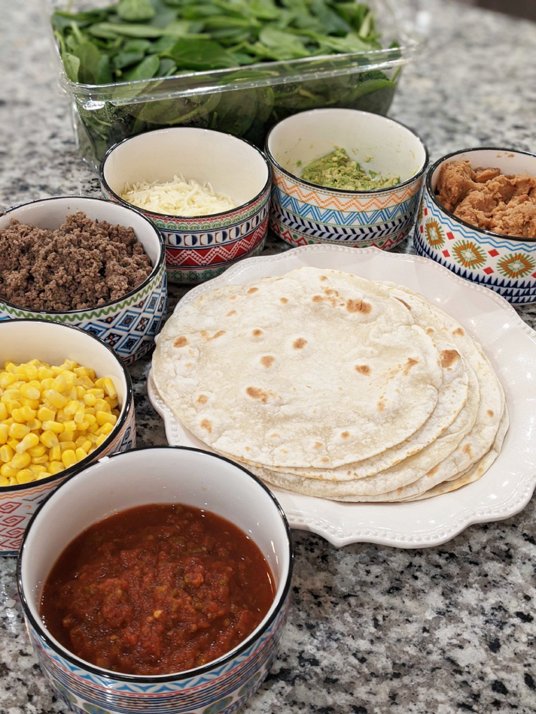 Taco bar with 6 bowls with taco fixings, and tortillas in the middle.